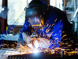 Welder working beneath a welding fume extraction system in an industrial fabrication facility while following workplace safety procedures.